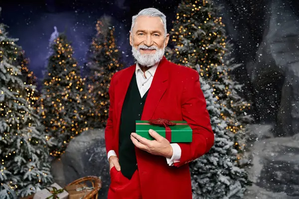 A smiling gentleman dressed in a vibrant red outfit presents a wrapped gift in a snowy setting. — Stock Photo