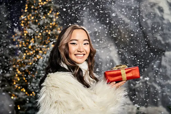 A joyful woman enjoys the festive spirit while holding a red present amidst falling snow. — Stock Photo