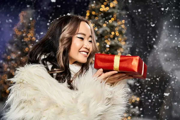 A young woman smiles brightly while cradling a red gift with gold ribbon among sparkling lights. — Stock Photo