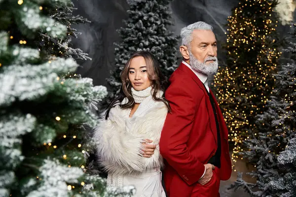 A woman in a white coat and a man in a red suit stand back to back surrounded by Christmas trees. — Stock Photo
