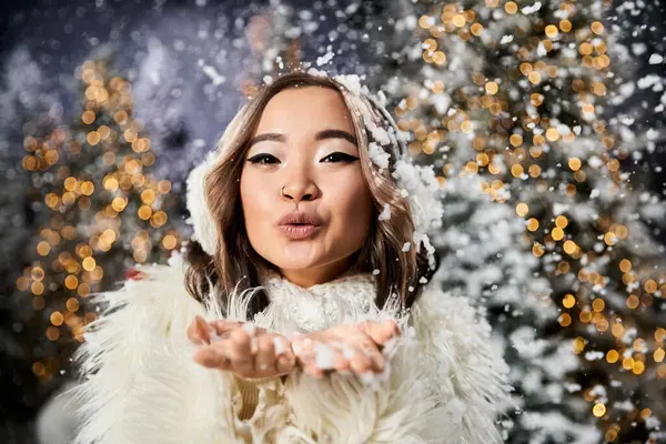 Une jeune femme en tenue d'hiver souffle ludique des flocons de neige, entourée de lumières de vacances éclatantes. — Photo de stock