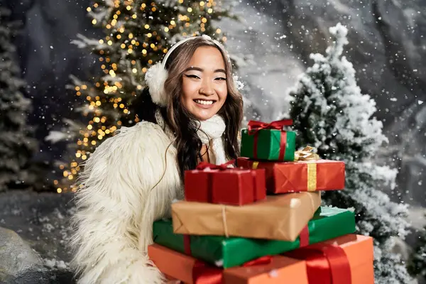 A cheerful young woman in a cozy coat sits with colorful gifts amid a snowy backdrop. — Stock Photo
