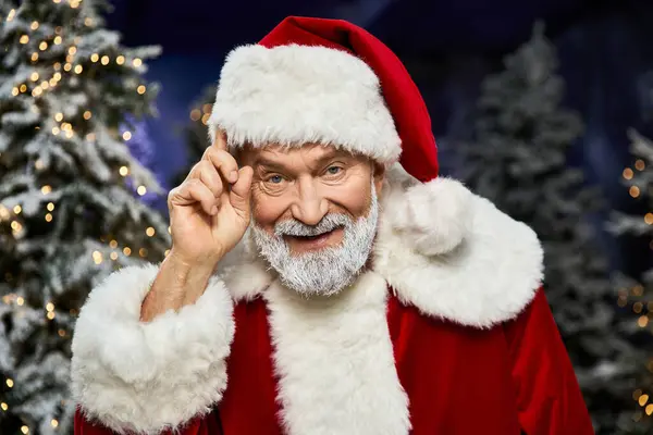 A cheerful Santa Claus tips his hat while surrounded by magical snow laden evergreen trees. — Stock Photo