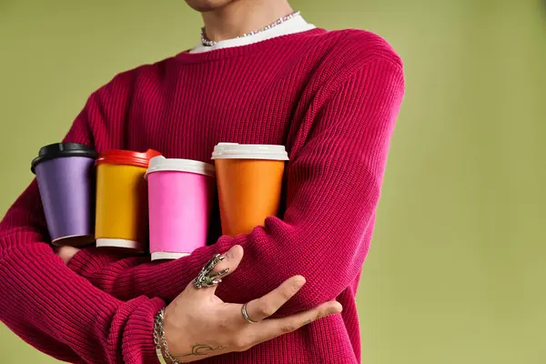 A fashionable young man stands confidently, cradling an array of vibrant coffee cups on a serene background. — Stock Photo