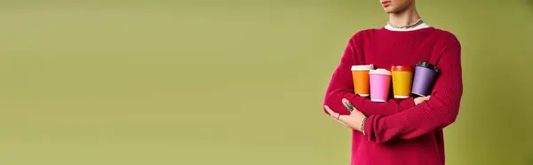 A fashionable young man wearing a pink sweater poses confidently, holding an array of vibrant coffee cups. — Stock Photo
