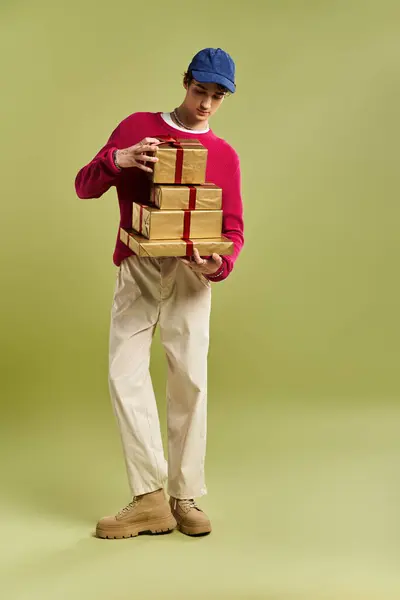 A stylish young man in a vibrant sweater joyfully presents beautifully wrapped Christmas gifts in a studio. — Stock Photo