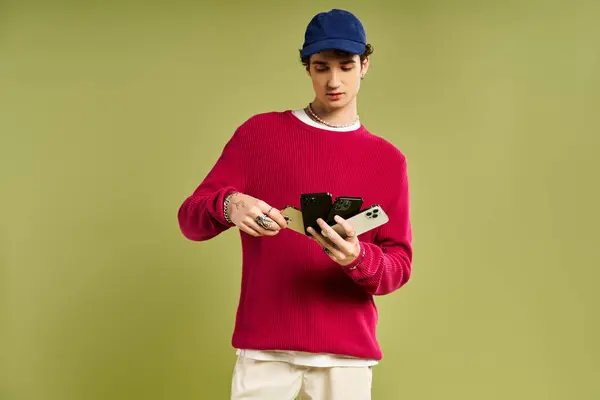 Ein stylischer junger Mann im rosafarbenen Pullover posiert selbstbewusst mit mehreren Smartphones vor grünem Hintergrund. — Stockfoto