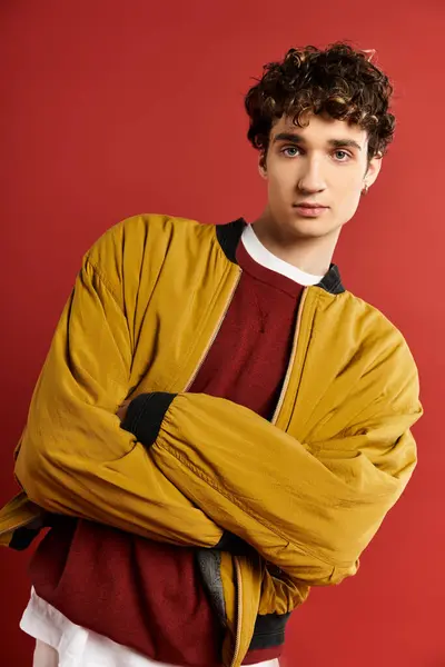 A handsome young man with curly hair stands with arms crossed, wearing a yellow jacket on a striking red backdrop. — Stock Photo