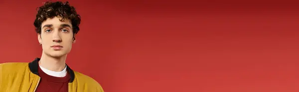 A young man with stylish curly hair showcases his yellow jacket while standing against a bold red wall. — Stock Photo