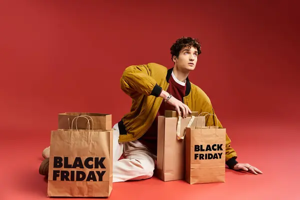 Un joven guapo sostiene con entusiasmo las bolsas de la compra mientras está sentado en un estudio con un audaz telón de fondo rojo. - foto de stock