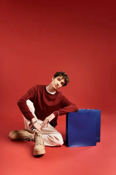 A handsome young man with curly hair sits casually, showcasing shopping bags amidst a bold backdrop. — Stock Photo