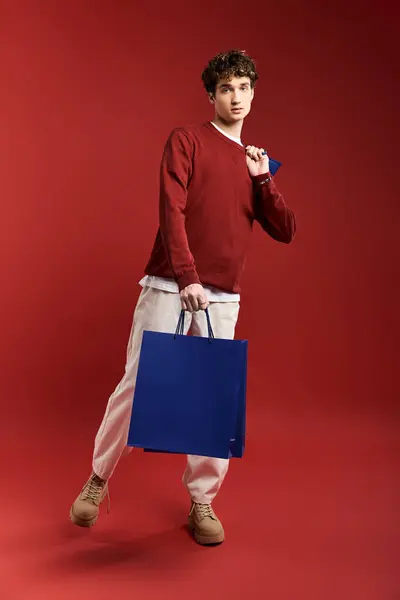 Ein stylischer junger Mann mit lockigem Haar hält bunte Einkaufstüten in der Hand und posiert selbstbewusst in einem Atelier. — Stockfoto