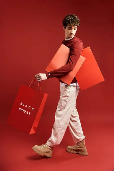 Un joven elegante con el pelo rizado lleva felizmente bolsas de compras en un vibrante estudio el Viernes Negro. - foto de stock