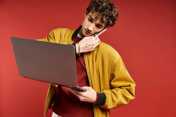 A fashionable young man in a yellow jacket balances a laptop and phone while posing against a vibrant red background. — Stock Photo