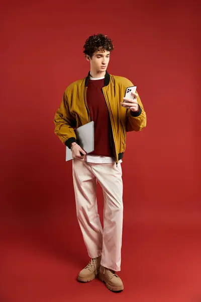 A handsome young man with curly hair holds a phone and tablet while posing in a yellow jacket on red. — Stock Photo