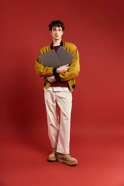 A handsome young man stands poised, cradling two laptops in his arms, dressed in a trendy yellow jacket. — Stock Photo
