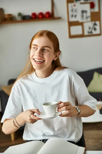 A cheerful young man sips his tea, immersed in studying at his warm and inviting workspace. — Stock Photo