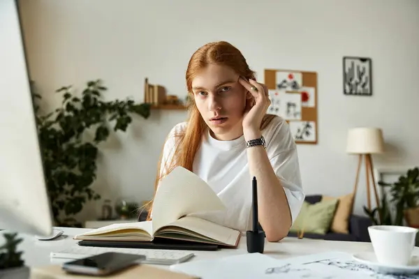 A young man with long hair looks thoughtfully at an open book in a sunny, inviting study. — Stock Photo