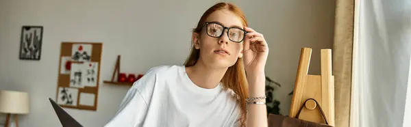 A young man with glasses sits thoughtfully, surrounded by artistic elements, lost in study. — Stock Photo