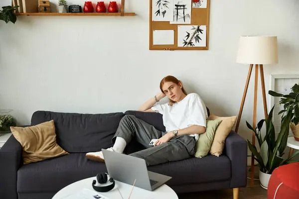 A young man in casual attire sits comfortably on a couch, working on a laptop in a bright space. — Stock Photo