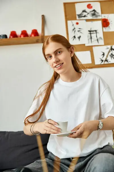 Le jeune homme tient une tasse de thé, souriant dans une chambre élégante et détendue ornée d'art. — Photo de stock