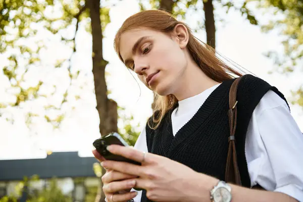 Ein junger Mann mit langen Haaren konzentriert sich auf sein Smartphone inmitten des üppigen Grüns im Park. — Stockfoto