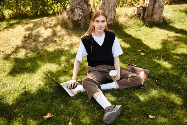 He is seated on the grass with a laptop and a warm drink, engaging with his work outdoors. — Stock Photo