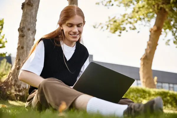 A young man sits on the grass, focused on his laptop, with a smile on his face. — Stock Photo