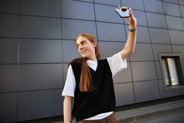 A young man with long hair smiles and waves while taking a selfie outdoors. — Stock Photo