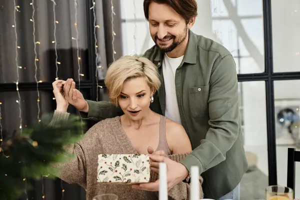 Una pareja celebra el amor en su acogedora casa, desenvolviendo un regalo sorpresa en medio de una cálida decoración. - foto de stock