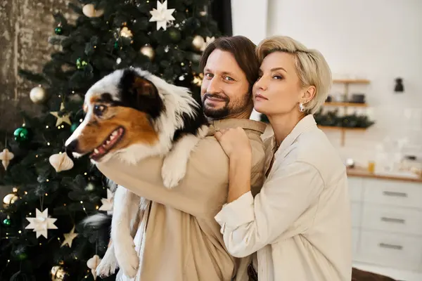 A happy couple holds their dog warmly at home, surrounded by festive holiday decorations. — Stock Photo