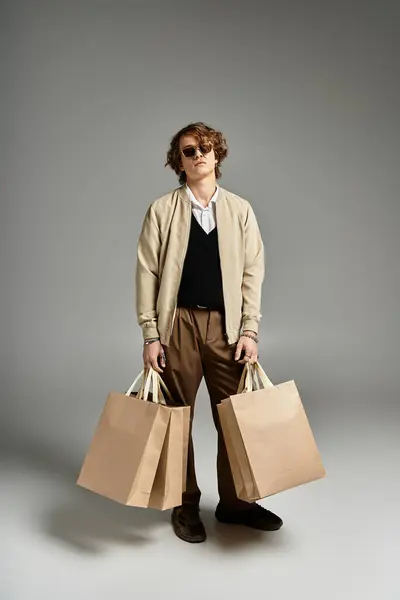 A dapper young man stands confidently with two shopping bags, showcasing elegant autumn attire in a studio. — Stock Photo