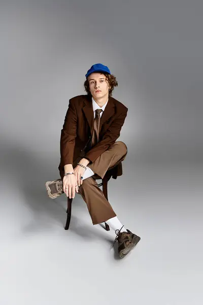 A confident young man sits elegantly on a chair, dressed in a brown suit and blue baseball cap, exuding charm. — Stock Photo