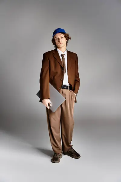 A young man stands poised in a brown suit and baseball cap, holding a laptop while showcasing his unique style. — Stock Photo