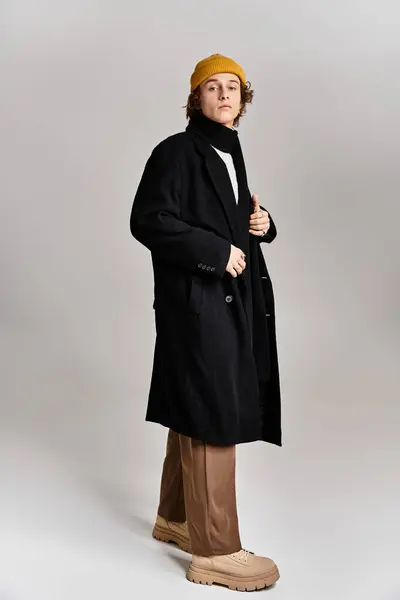 Un giovane con un cappotto nero e un berretto giallo mette in mostra la moda moderna in un elegante ambiente da studio. — Foto stock