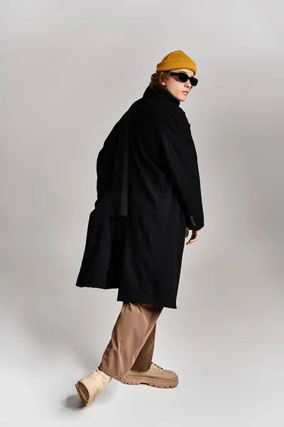 A handsome man in a black coat and yellow beanie stands poised in a modern studio, exuding cool confidence. — Stock Photo