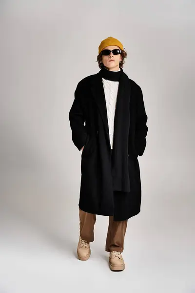 The confident young man stands tall in a chic black coat with a vibrant yellow beanie, showcasing urban style. — Stock Photo