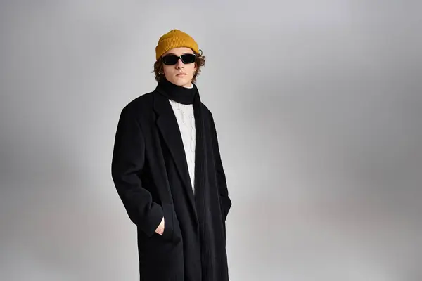 Un giovane alla moda mette in mostra il suo stile con un berretto giallo e un cappotto nero, trasudando fiducia in uno studio. — Stock Photo