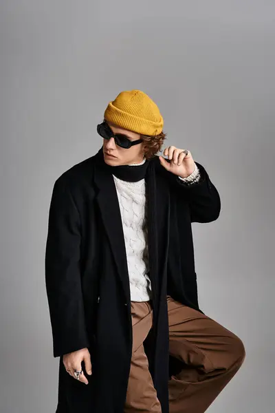 A handsome young man showcases his unique style with a black coat and a vibrant yellow beanie in a studio. — Stock Photo