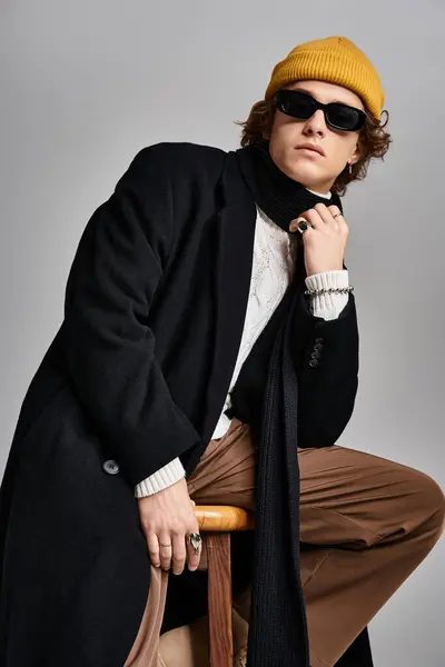 Un joven de moda con un abrigo negro y gorro amarillo emana confianza mientras está sentado en un estudio. - foto de stock