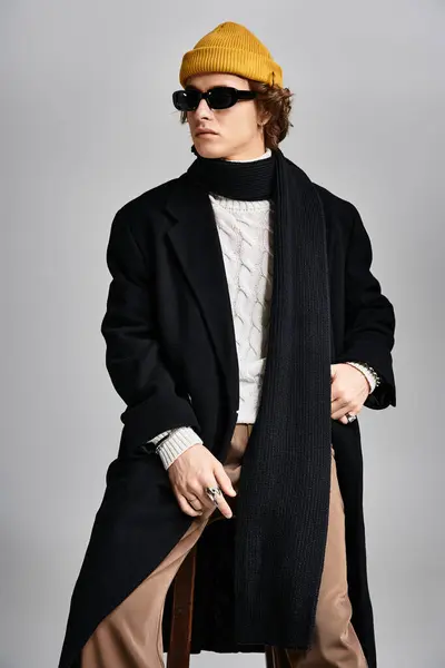 Un joven guapo está de pie con confianza, mostrando un abrigo negro de moda y un gorro amarillo vibrante en el estudio. - foto de stock