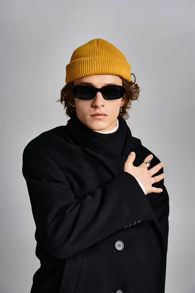 Un giovane alla moda mette in mostra il suo stile unico con un cappotto nero e un vivace berretto giallo in uno studio. — Foto stock