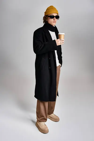 Un bel giovane uomo tiene una tazza di caffè mentre mostra il suo cappotto nero alla moda e il berretto giallo in uno studio. — Foto stock