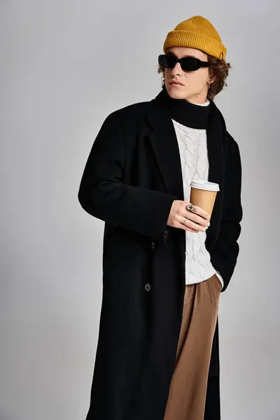 Un giovane fiducioso in un cappotto nero chic e un berretto giallo brillante tiene una tazza di caffè, radiante stile. — Foto stock