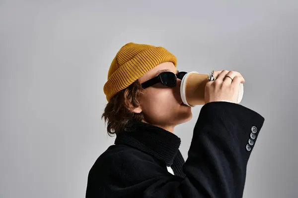 Un bel giovanotto sorseggia caffè mentre indossa un cappotto nero alla moda e un cappello giallo brillante. — Foto stock