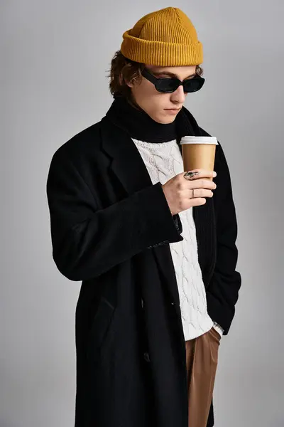 Un giovane alla moda si alza con fiducia, godendo il suo caffè mentre vestito con un cappotto nero e un berretto vibrante. — Foto stock
