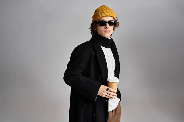 Un bel giovane con un cappotto nero e un berretto giallo si erge con fiducia con una tazza di caffè in mano. — Foto stock