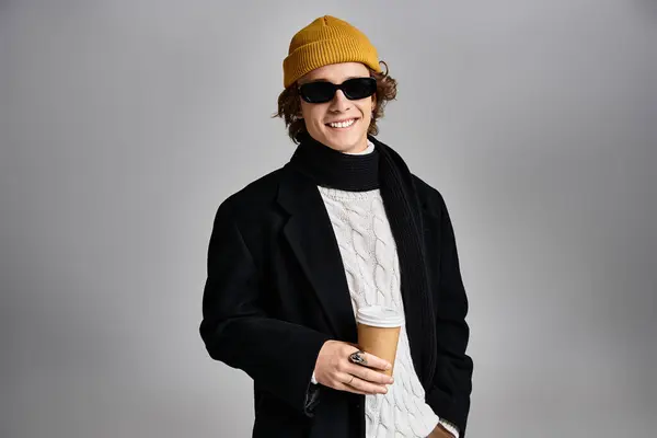 Un giovane uomo vestito con un cappotto nero e berretto giallo tiene una tazza di caffè, trasudando stile in un ambiente contemporaneo. — Foto stock