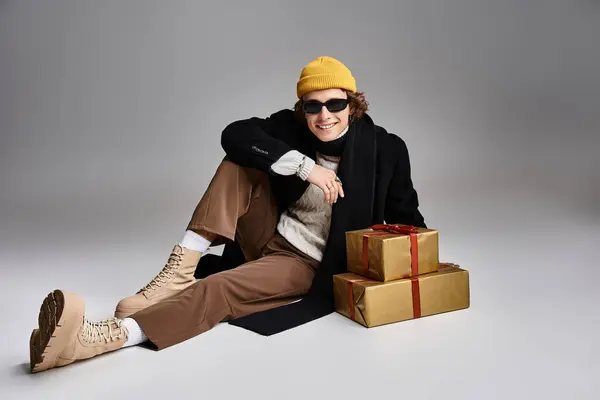 Un giovane alla moda, vestito con un cappotto nero e un berretto giallo, siede accanto a regali ben confezionati.. — Foto stock