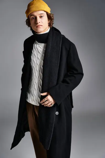 A fashionable young man wears a black coat and yellow beanie, showcasing a confident pose in a studio. — Stock Photo